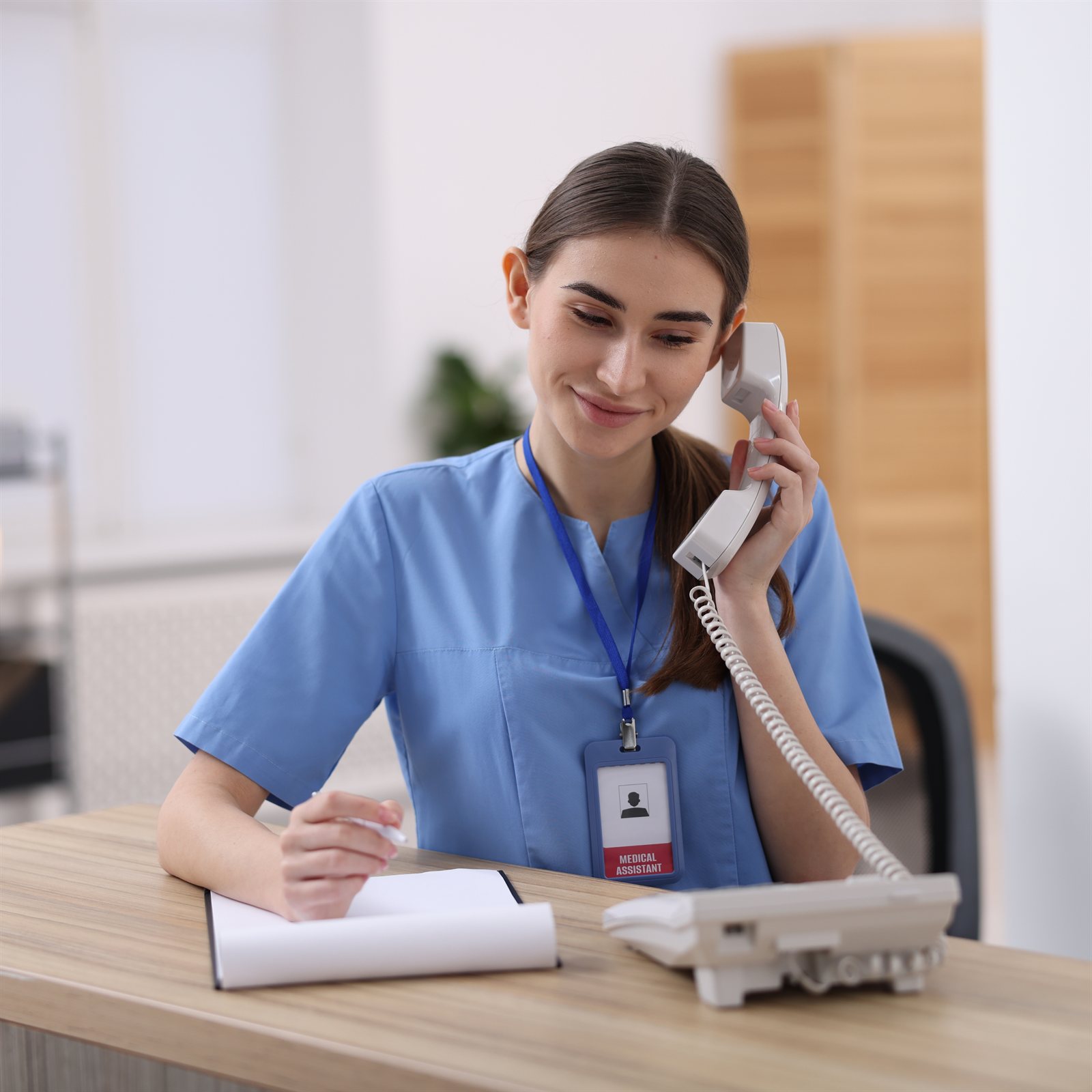 Profissional de saúde na recepção de uma clínica, atendendo ao telefone e anotando o agendamento.